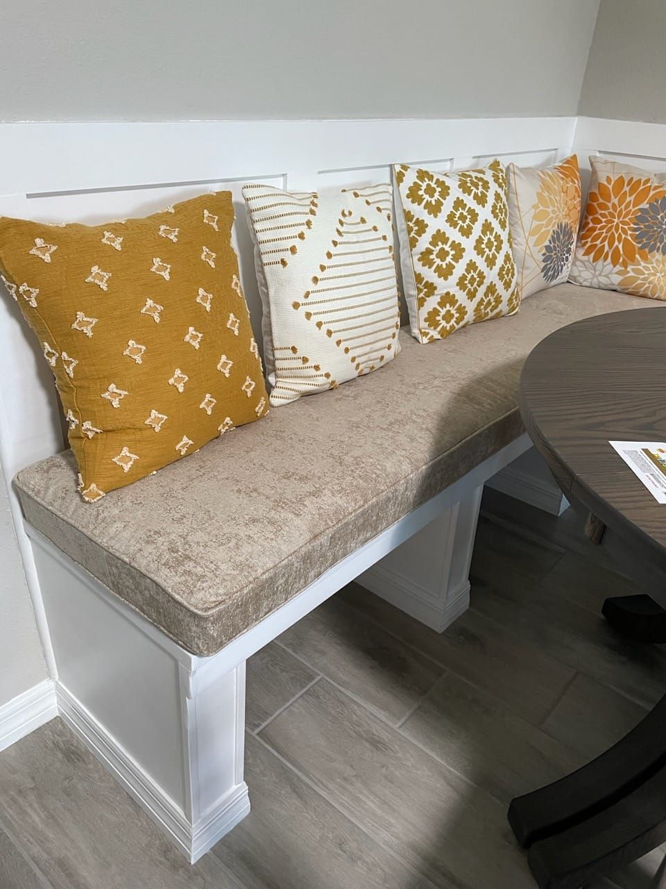 Built-in dining bench with tan cushion and decorative pillows in yellow and white, next to a round table.
