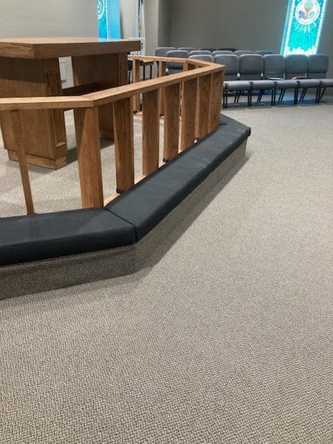 A church interior with wooden railing and cushioned bench. Gray carpet and chairs are visible.