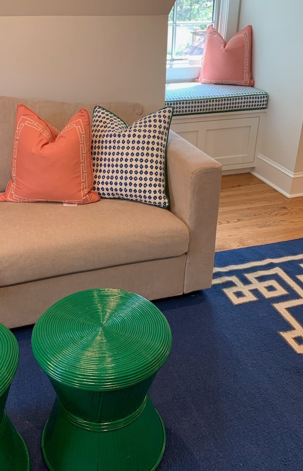 Beige couch with coral and blue pillows, window seat, green stools, and blue rug.