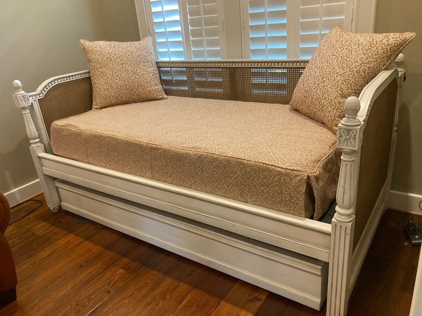 White daybed with woven back and patterned cushions, in front of shuttered window.