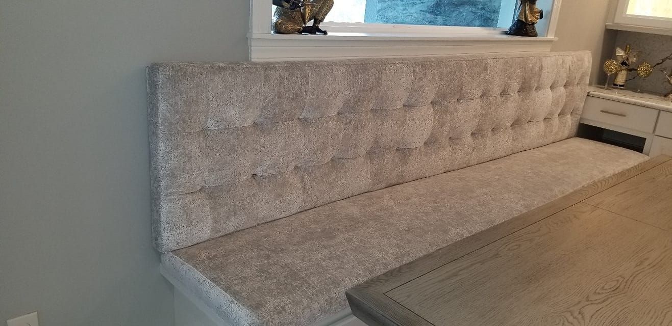 Built-in, tufted bench with a light-colored fabric against a gray wall and a table.