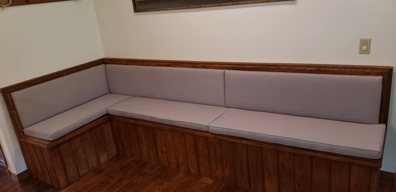 Built-in, L-shaped wooden bench with gray cushions in a corner; light walls and a wooden floor.