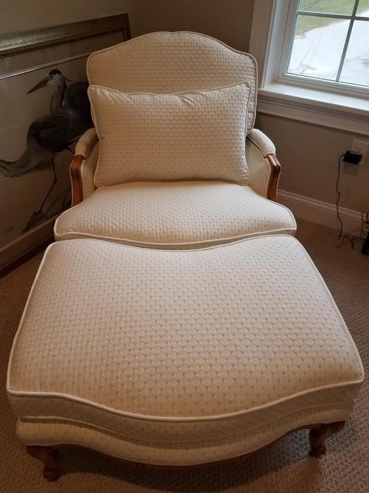 Cream-colored upholstered chaise lounge with matching footrest, wooden frame and trim, indoor setting.
