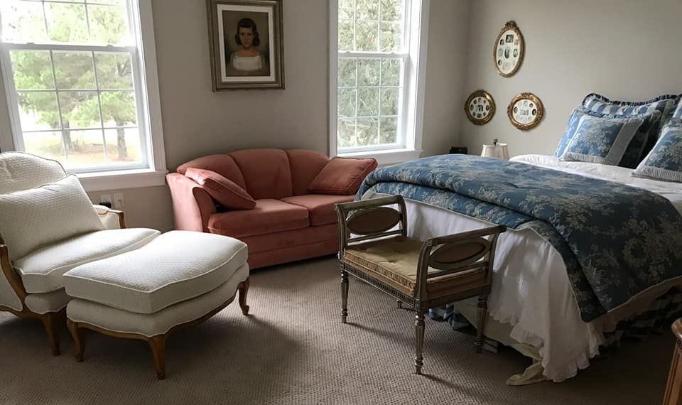 Bedroom with a bed, seating area with a sofa, chair, and a bench. Light walls and windows; neutral carpet.