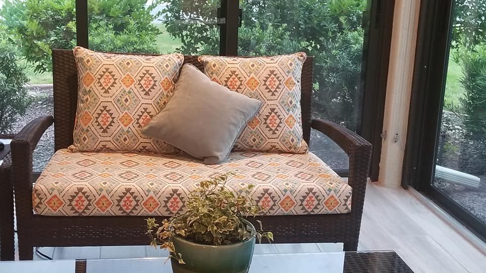 A wicker loveseat with patterned cushions and a gray pillow, beside a glass window, with a small potted plant in front.