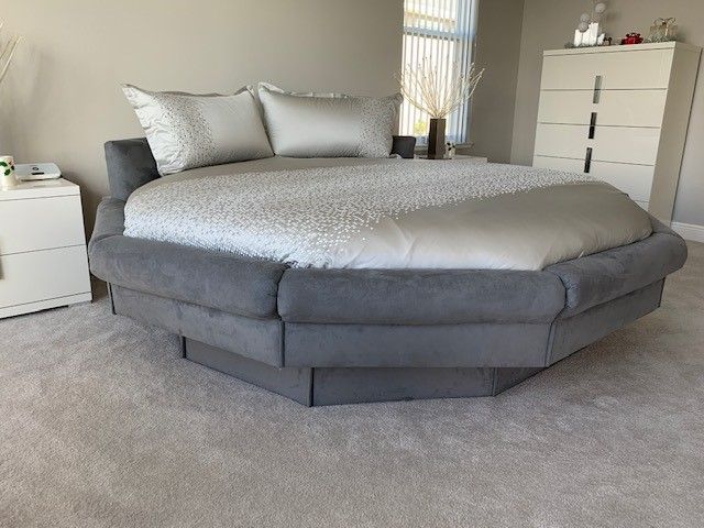 A large, round bed with gray upholstery and silver bedding sits in a bedroom with neutral-colored walls and carpet.