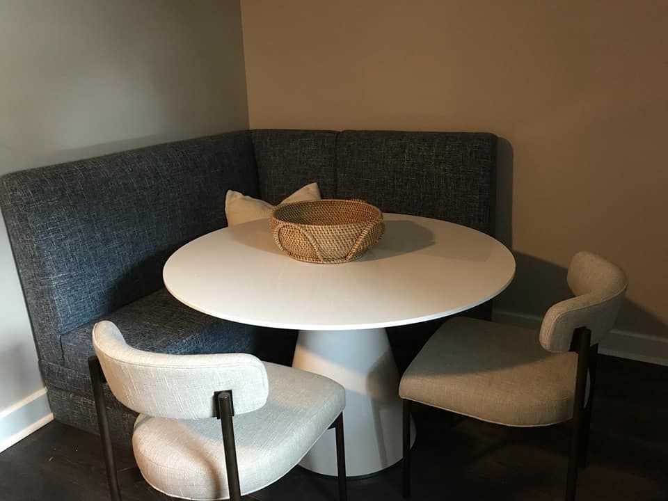 A cozy dining nook features a round white table with a woven bowl centerpiece, a built-in blue upholstered bench, and two cream-colored chairs.
