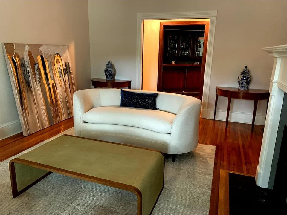 A cozy living room with a white curved sofa, coffee table, and abstract art. Dark wood floors and two antique tables flank a doorway.