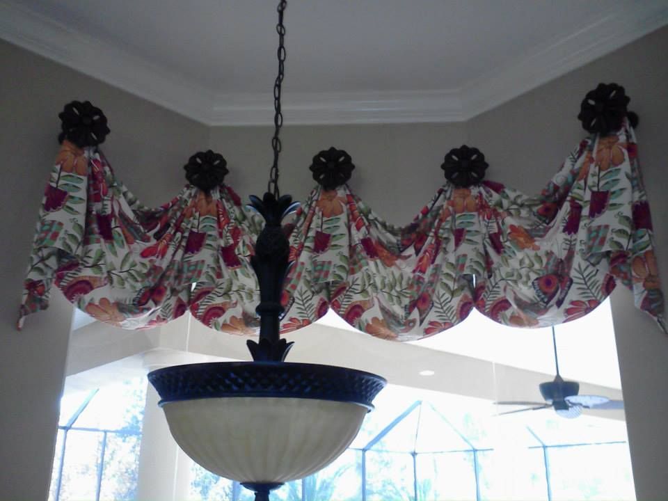 Floral fabric valance draped along a beige wall, above a hanging chandelier, in a room with a high ceiling.