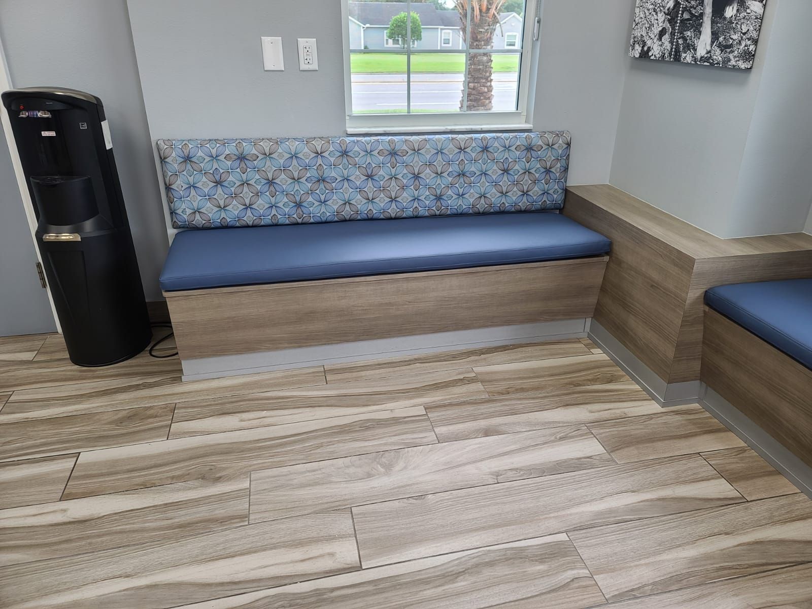 Waiting area with a blue and patterned bench seat, wooden-look cabinets, and a water cooler. The setting includes a window and framed artwork.