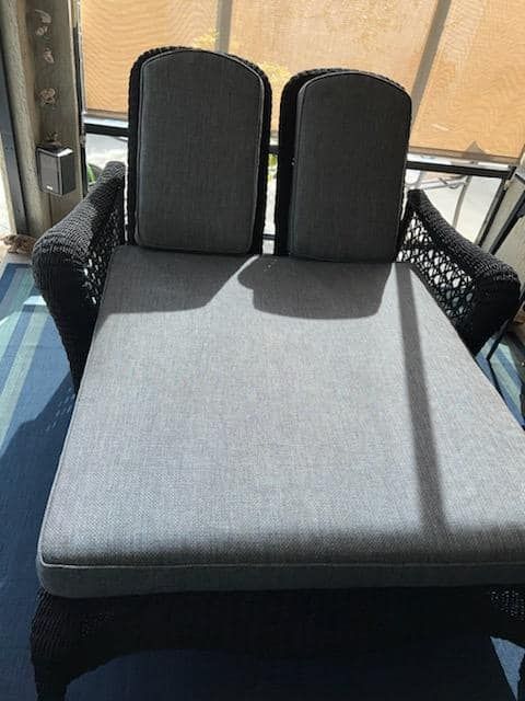 A dark wicker outdoor chaise lounge with gray cushions sits in a sunroom.