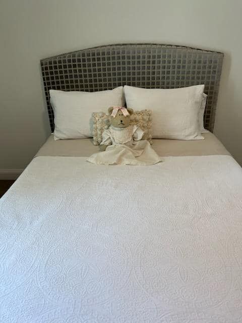 A bed with a white patterned quilt, two white pillows, and a teddy bear with angel wings. The headboard is gray with a grid pattern.