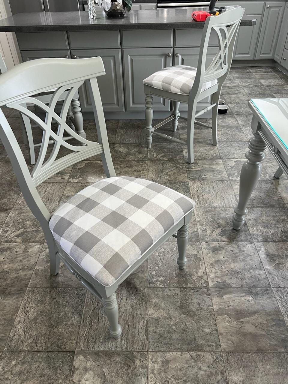 Two gray chairs with white and gray gingham cushions in a kitchen. The chairs are next to a table and a countertop with cabinets.