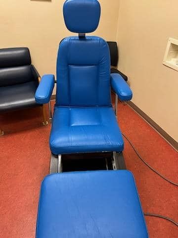 A blue medical examination chair in a room with a red floor. Another chair is visible in the background.
