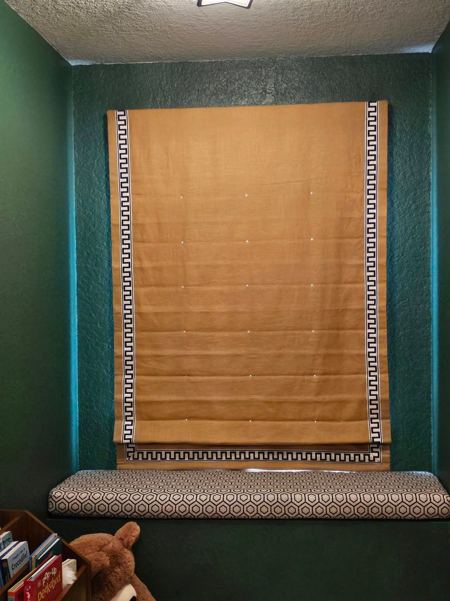 Tan Roman shade in front of a green wall, with a patterned bench below. Sides of the shade have a black and white design.