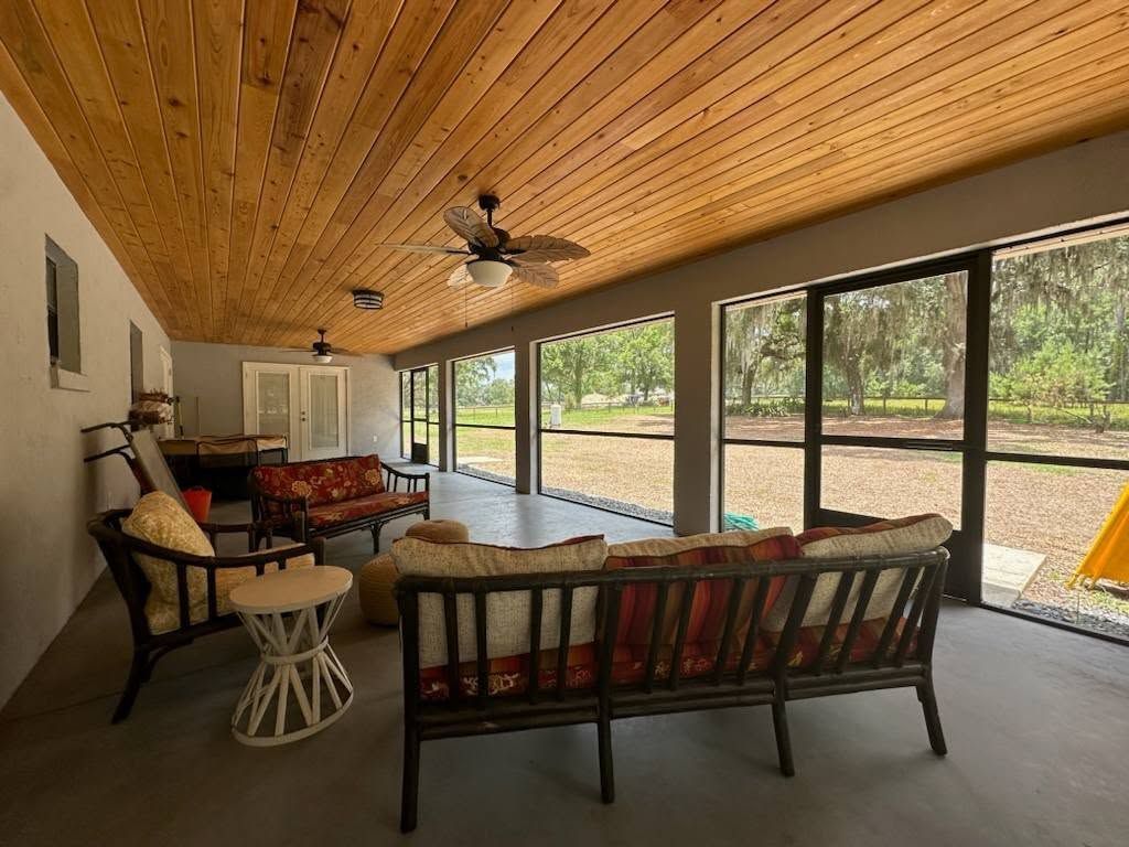 Covered patio with wooden ceiling, furnished with sofas and tables. Screens offer a view of green trees and grass.