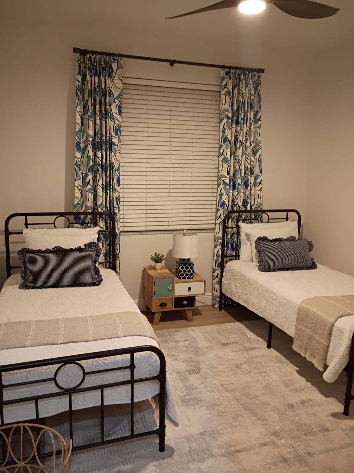 Two twin beds in a bedroom, with blue and white floral curtains and a light-colored rug. Black bed frames and nightstand.