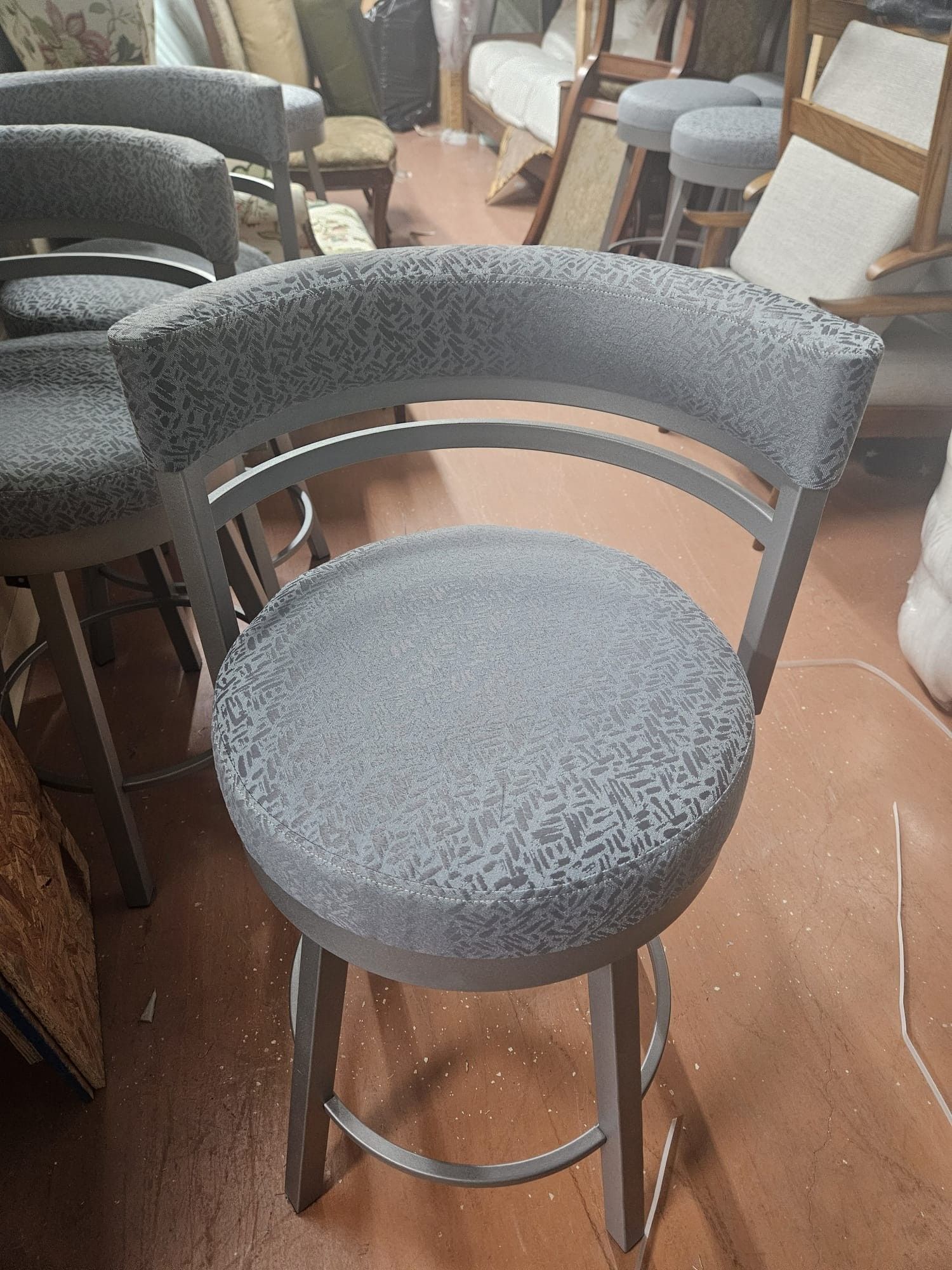 Gray upholstered bar stool in a workshop setting. The stool has a curved back and a round seat.