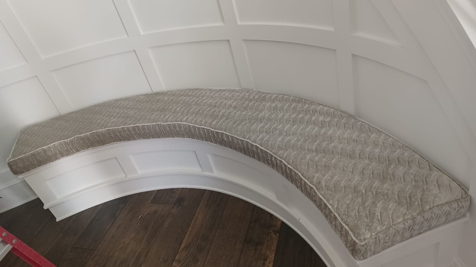 A curved built-in bench with a gray cushion and white trim sits against a curved wall. The bench is above a dark wood floor.