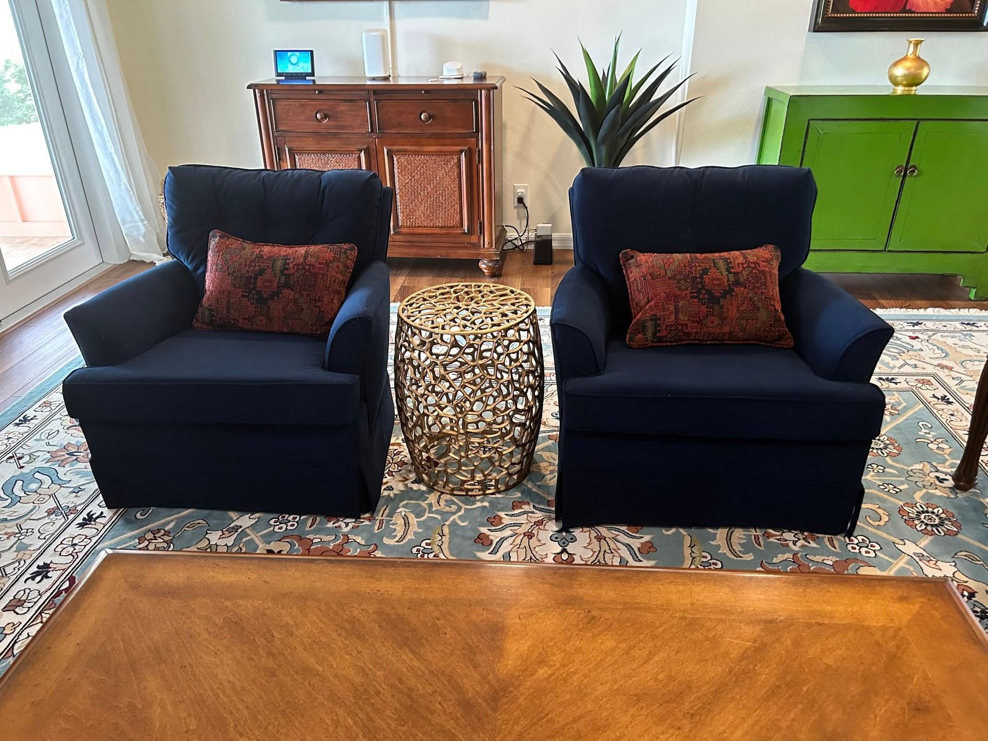 Two navy blue armchairs with patterned pillows face a gold side table, set on a patterned rug. In the background, there's a wooden cabinet and green credenza.