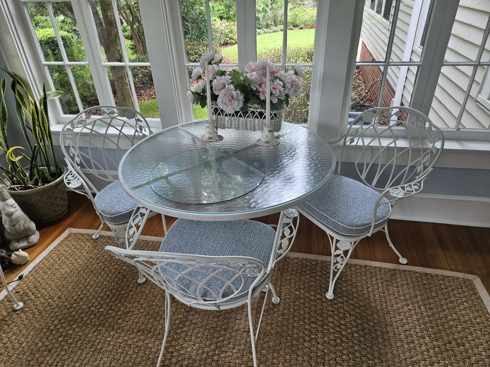 A sunroom with white wrought iron table and chairs, topped with a floral arrangement, and a patterned rug.