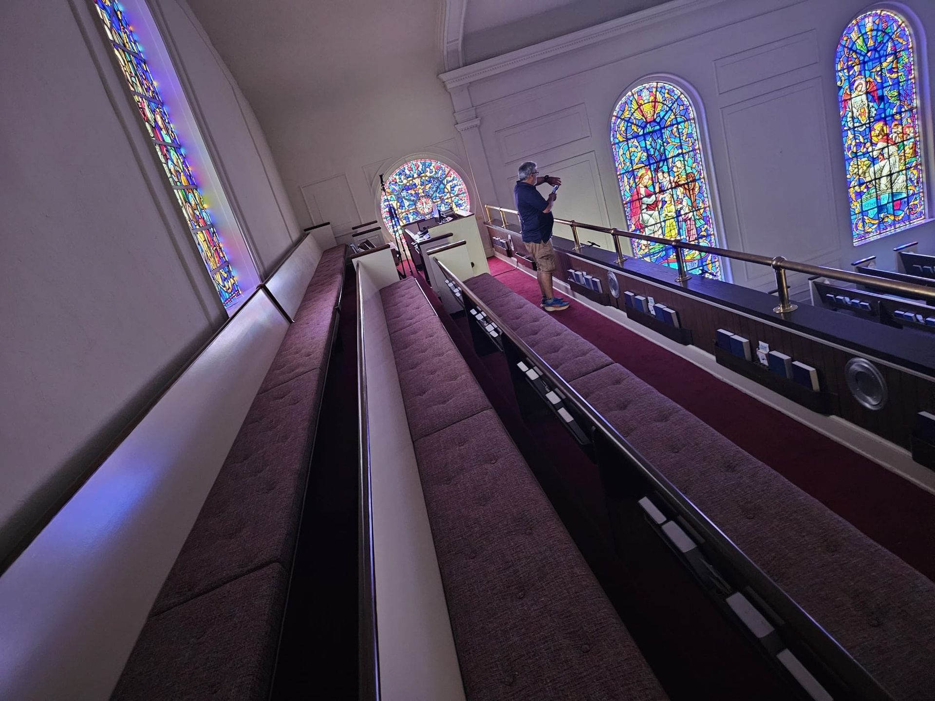 Inside a church, a person stands near the pews.  Stained glass windows line the walls.  Carpeting and a railing are also visible.