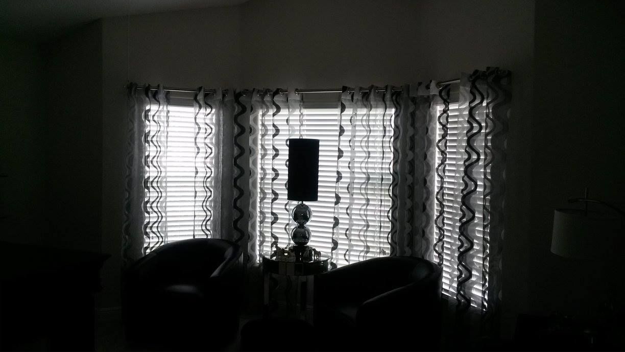 A room with large windows, wooden blinds, and patterned curtains. Two dark chairs face the windows with a lamp in front.