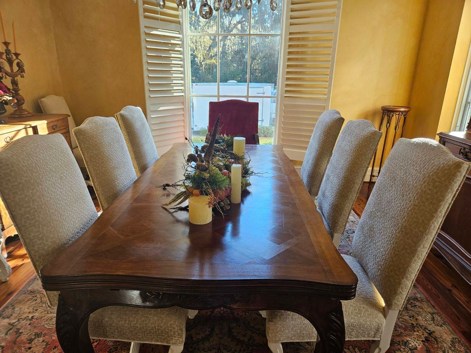 An elegant dining room with a long wooden table set for eight. The chairs are upholstered in neutral fabric, and the room has a large window with shutters.