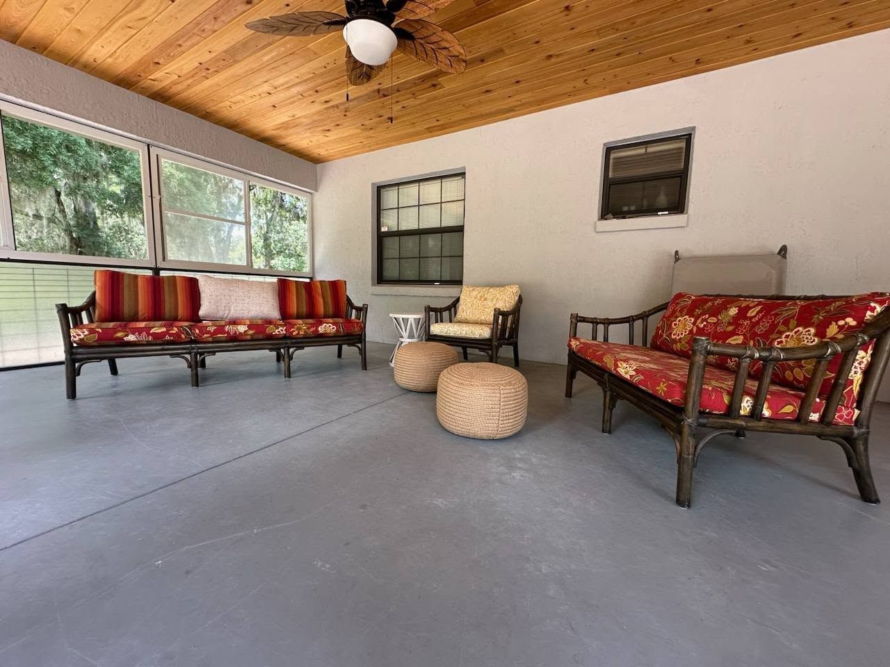 A covered porch with gray flooring and wicker furniture with red floral cushions. Two woven ottomans sit in the center.