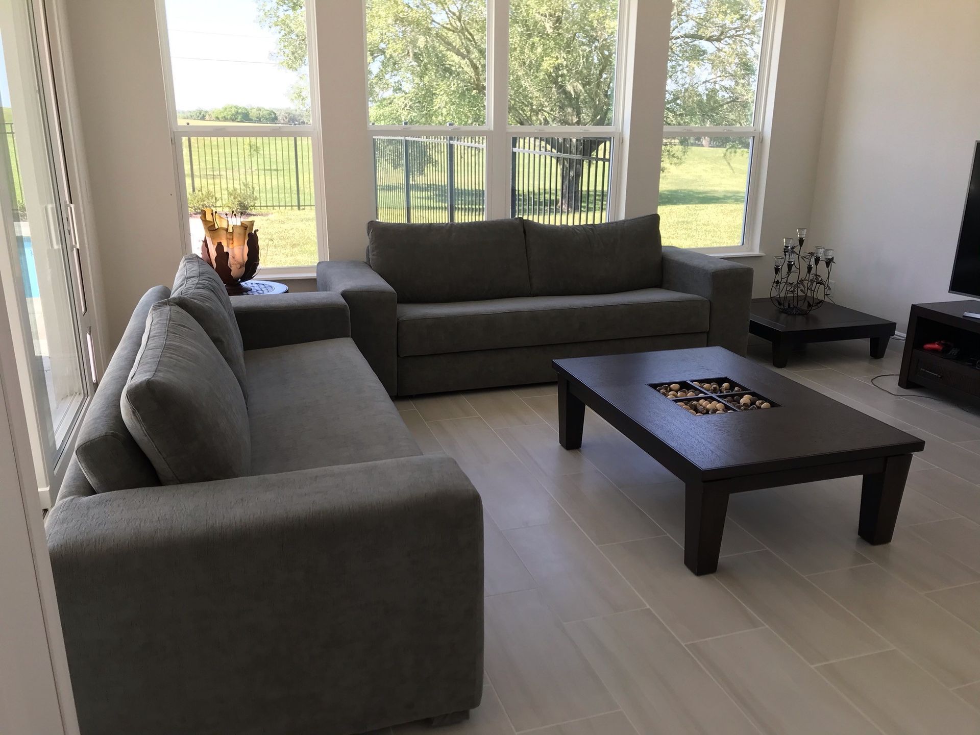 Living room with gray sofas, dark coffee table, and large windows overlooking greenery. Sunlight streams in.