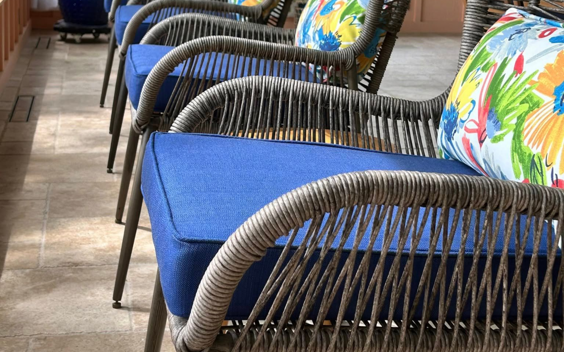 Row of woven gray chairs with blue cushions and floral pillows on a tiled patio.