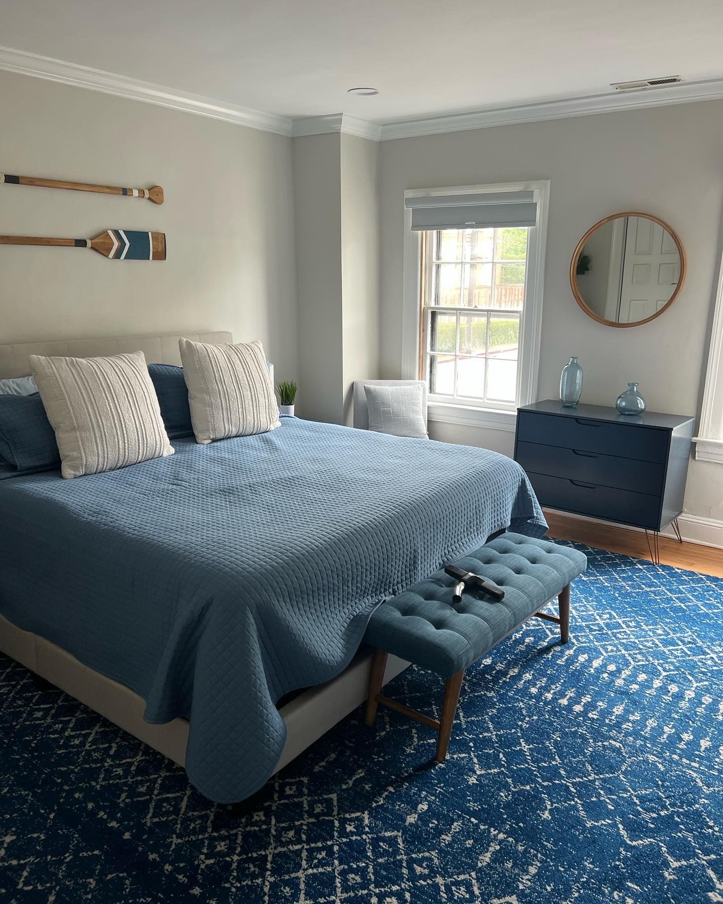 Blue bedroom with gray bed, patterned rug, dresser, round mirror, and window light