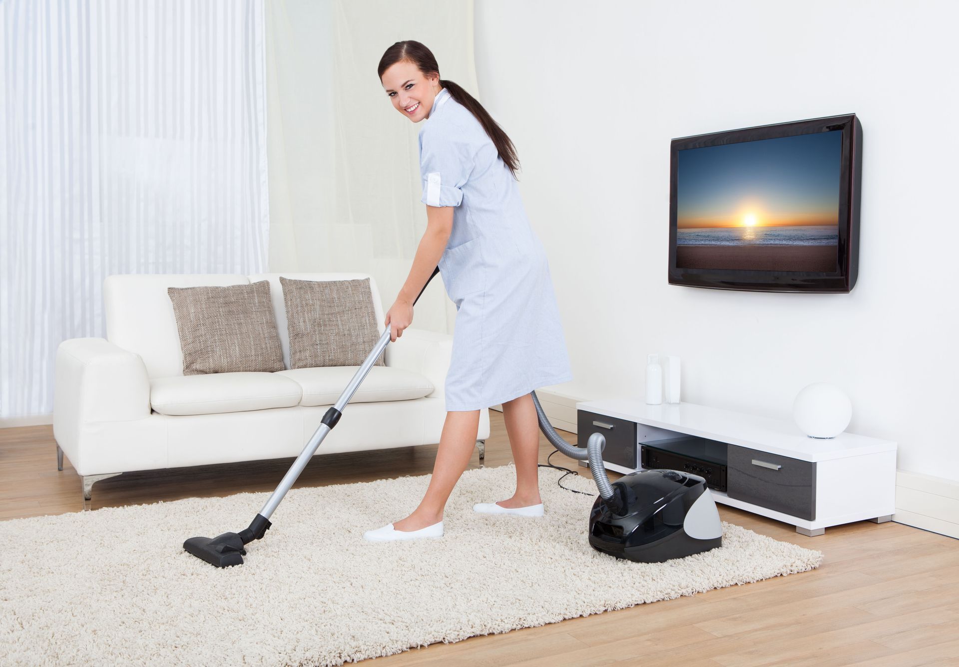 Person vacuuming a white rug in a bright living room with a sofa and wall-mounted TV.