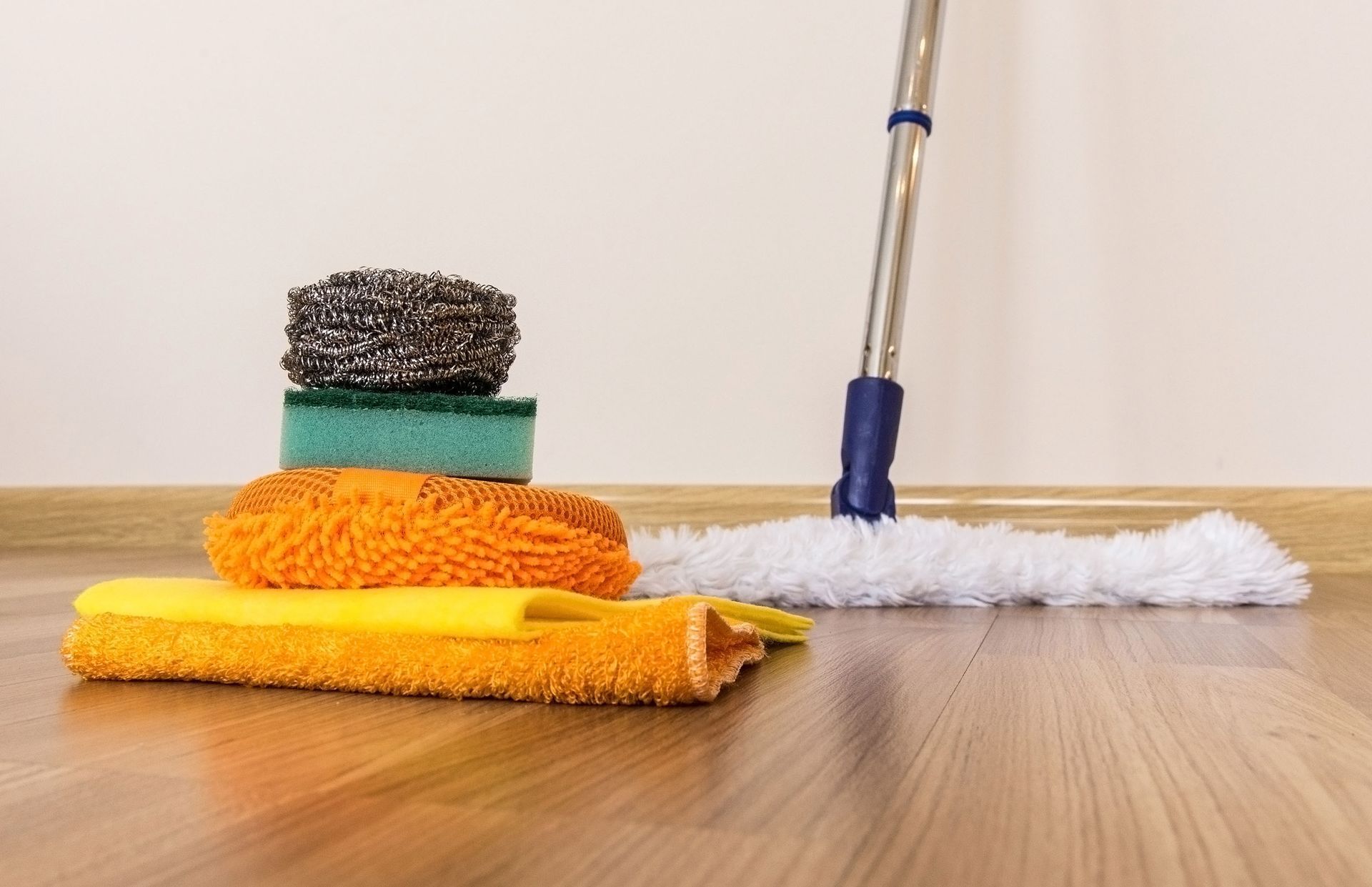 Cleaning supplies and mop on a wooden floor, with stacked scouring pads and sponges in the foreground