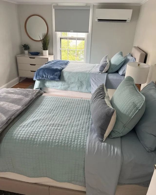 Bright bedroom with two neatly made twin beds, blue bedding, dresser, mirror, and window air conditioner.