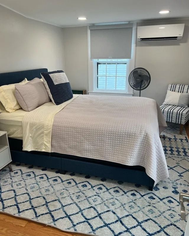 Bright bedroom with bed, patterned rug, window, fan, and wall-mounted air conditioner