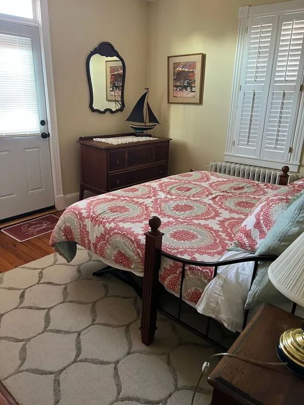 Neatly made bedroom with a patterned bedspread, dresser, mirror, window, and shuttered closet doors.
