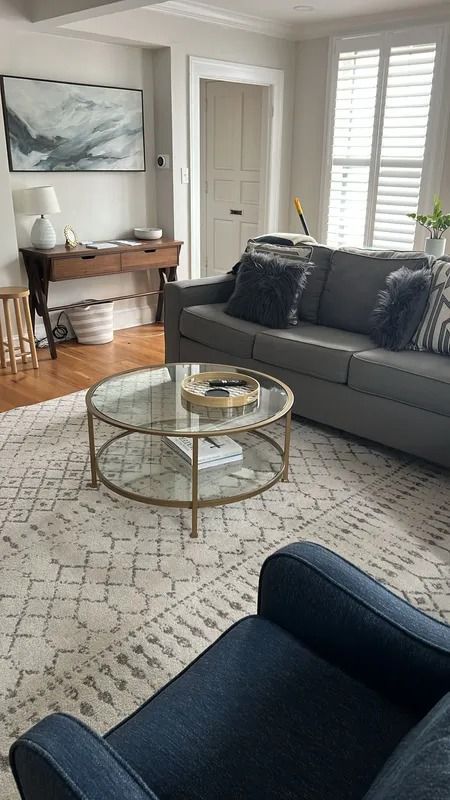 Bright living room with gray sofa, glass coffee table, navy chair, wooden desk, and window shutters