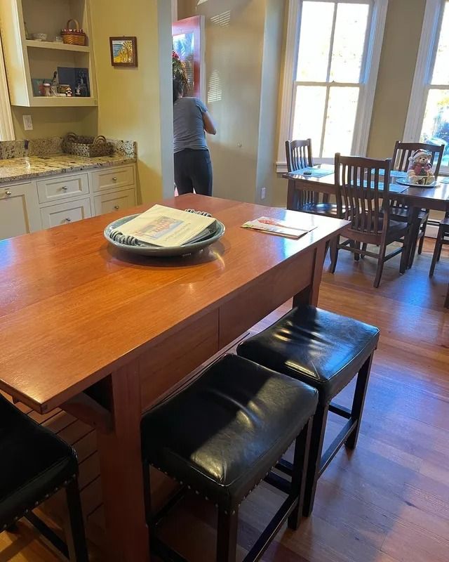 Kitchen with wooden island and stools; person standing near a doorway by a bright dining area.