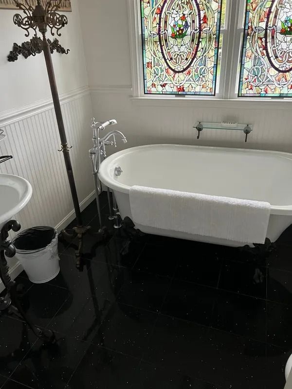 Small bathroom with a freestanding white bathtub, stained-glass window, sink, and black floor.