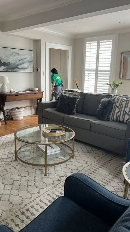 Living room with gray sofa, round glass coffee table, and a person cleaning near the window.