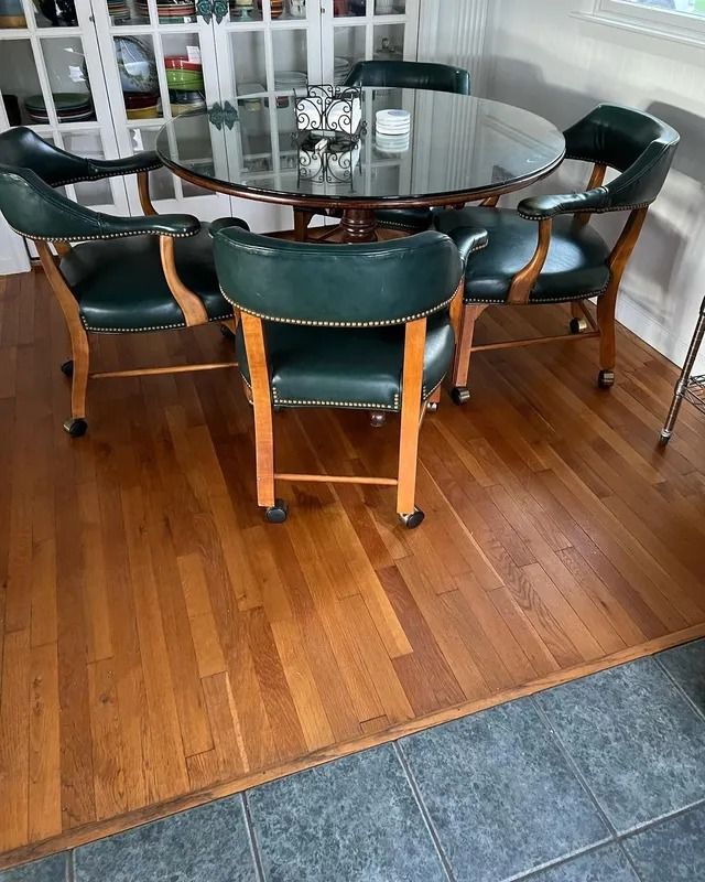 Round glass dining table with six dark green chairs on hardwood floor by a tiled entryway