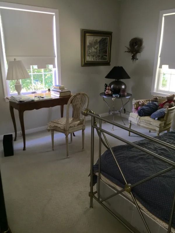 Bedroom with a metal bed, chair, table lamp, and two windows with blinds.