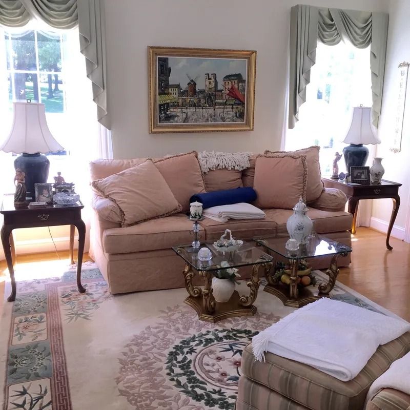 Elegant living room with beige sofa, glass coffee table, floral rug, and bright windows with drapes