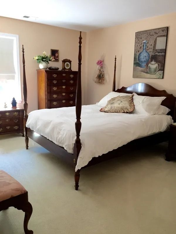 Spacious bedroom with four-poster bed, white bedding, dresser, and wall art in soft beige tones