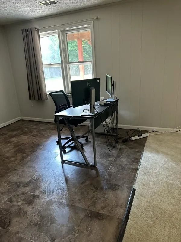 Empty room with a desk, monitor, chair, and large window on a bare floor.