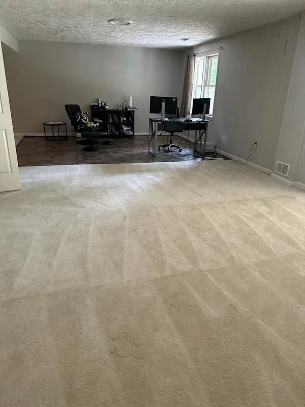 Empty beige carpeted living room opening to a dining area with dark wood floors and a desk by a window