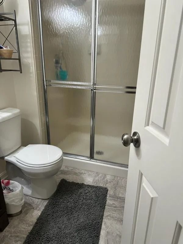 Small bathroom with a toilet, shower stall, dark bath mat, and open white door.