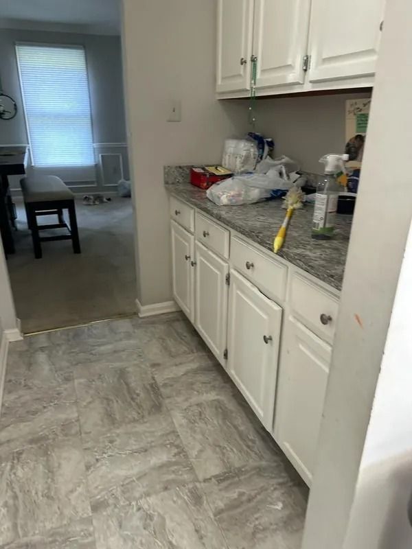 Kitchen with white cabinets and gray tile floor, cluttered countertop, leading into a dining area with a window.