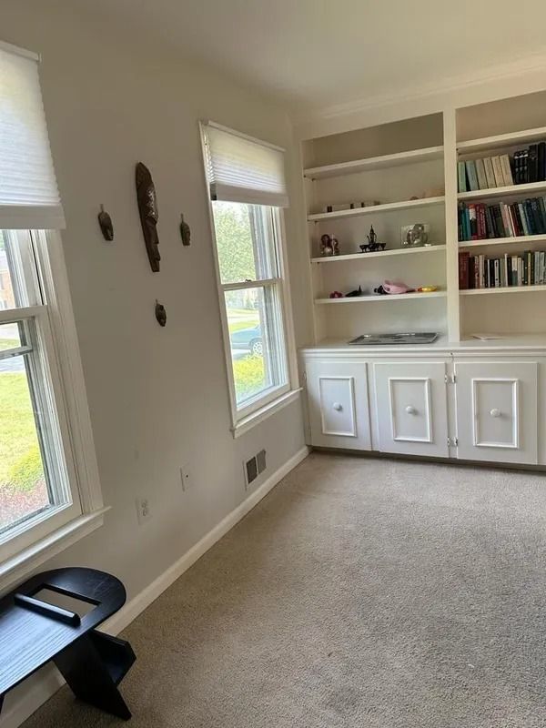 Small beige home office with built-in shelves, two windows, carpet, and a black bench near the wall.